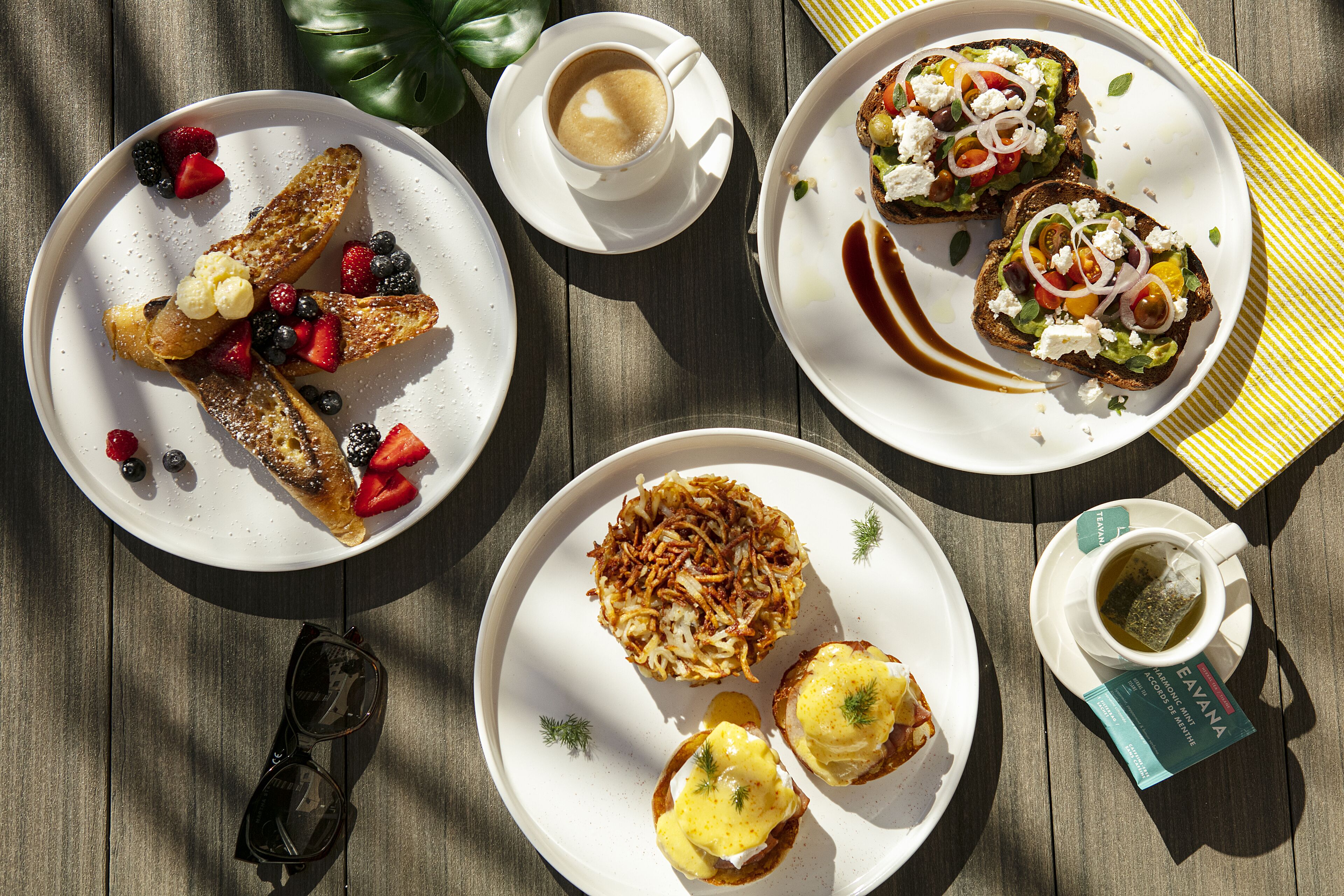 Une table dressée pour le petit-déjeuner, avec différents plats, notamment des toasts aux fruits rouges, des œufs Bénédicte, des pommes de terre rissolées et une tasse de thé.