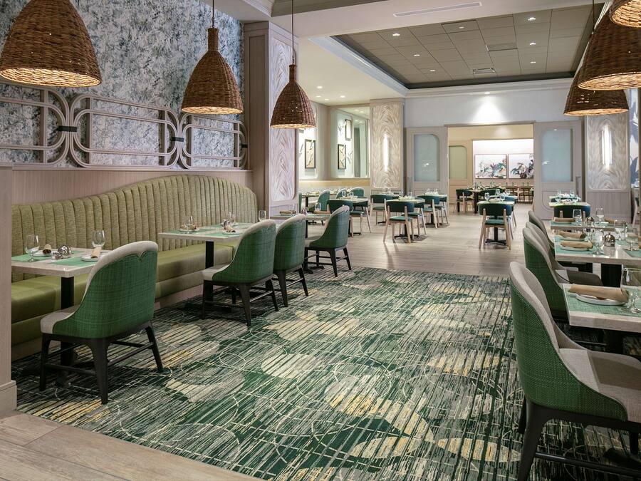 A restaurant with green upholstered booths and tables, wicker pendant lighting, and a patterned carpet.