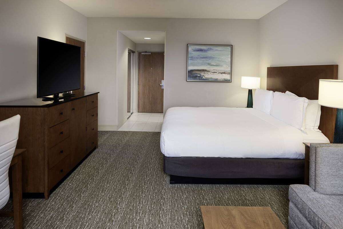 A hotel room with a large bed, a desk with a flat-screen TV, a chair, a lamp, and a painting on the wall.