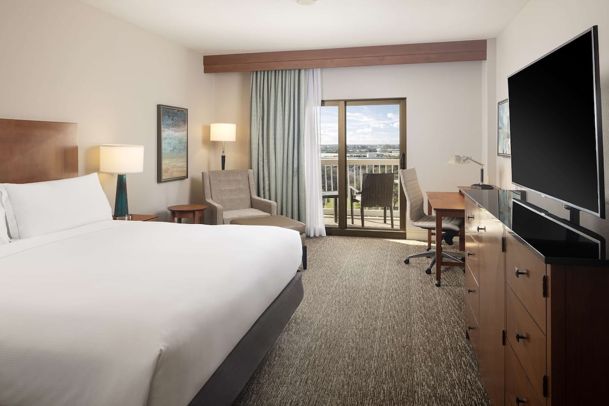 A hotel room with a large bed, a desk, a chair, a TV, and a balcony with a view.