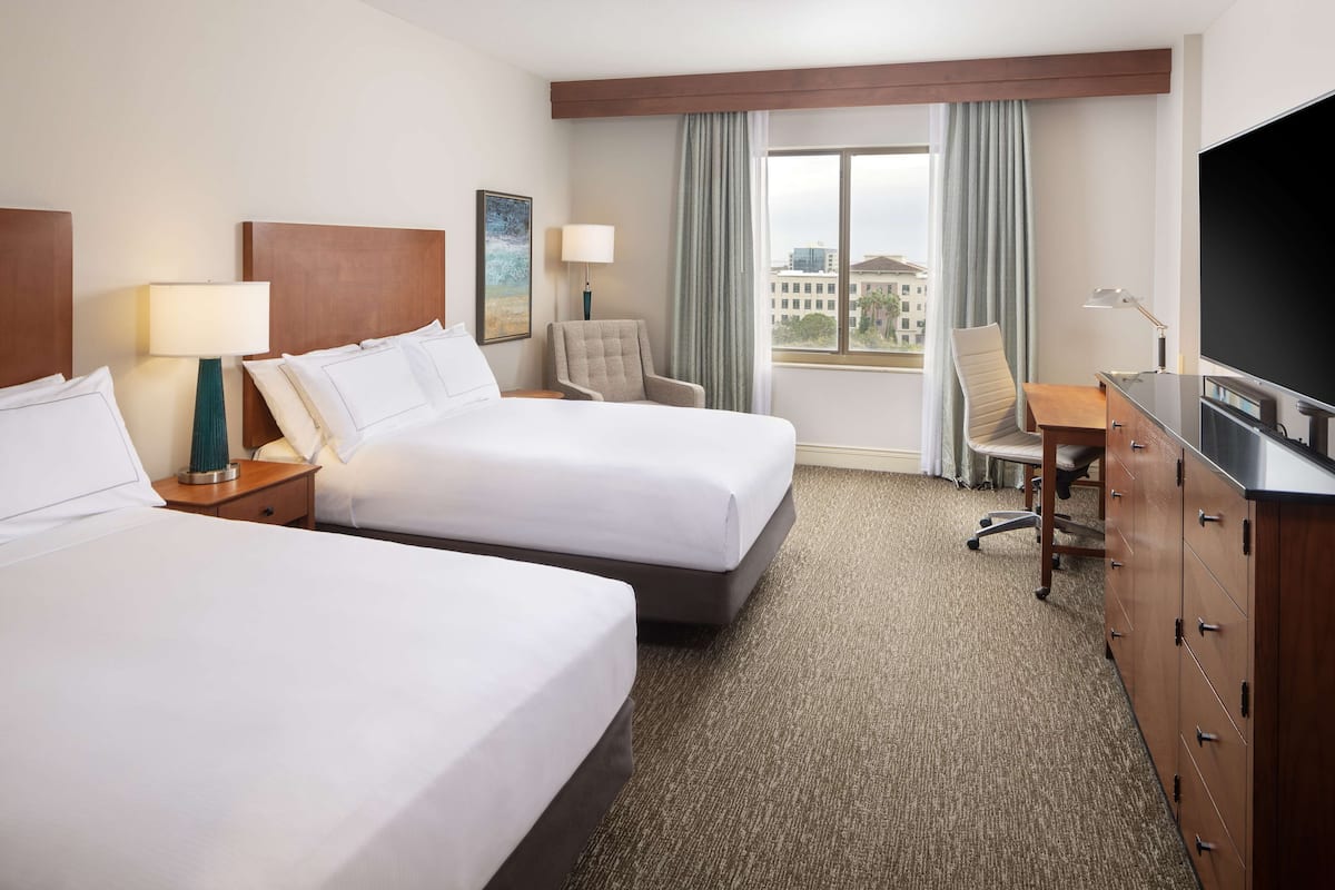 A hotel room with two beds, a desk, a TV, and a window with curtains.