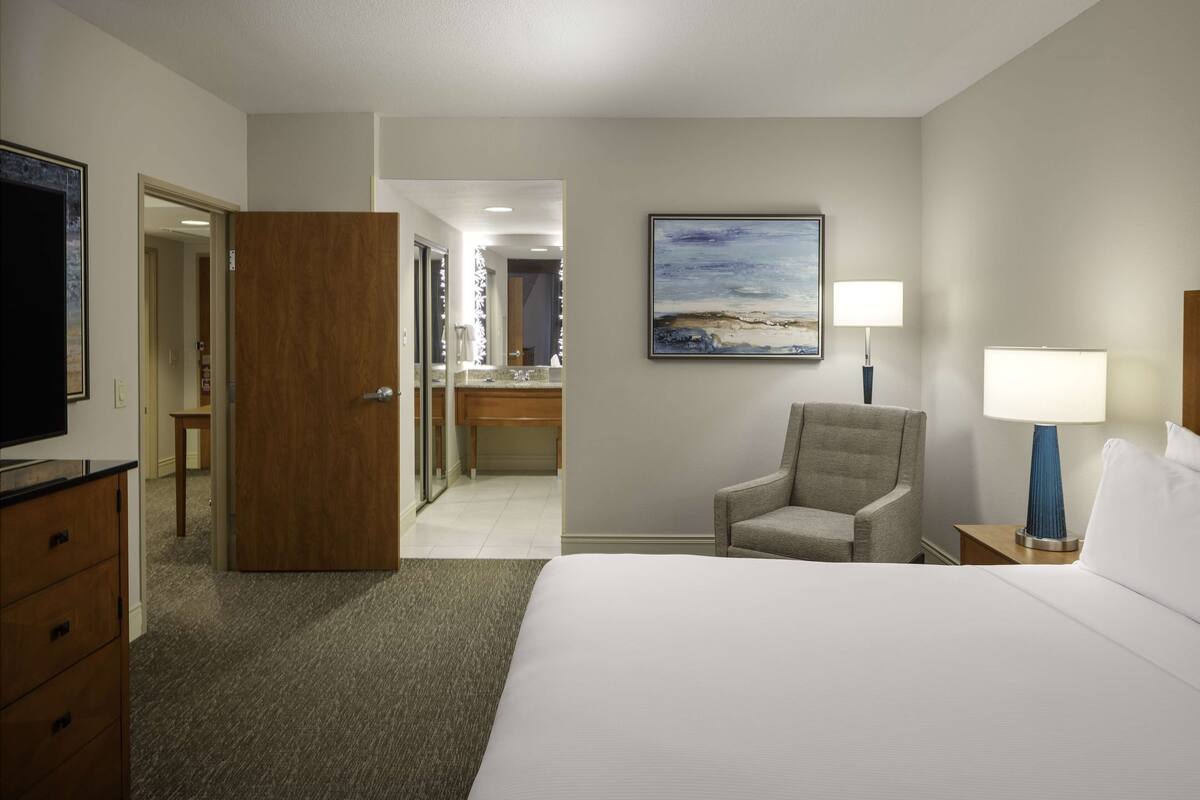 A hotel room with a bed, a chair, a desk, and a painting on the wall.