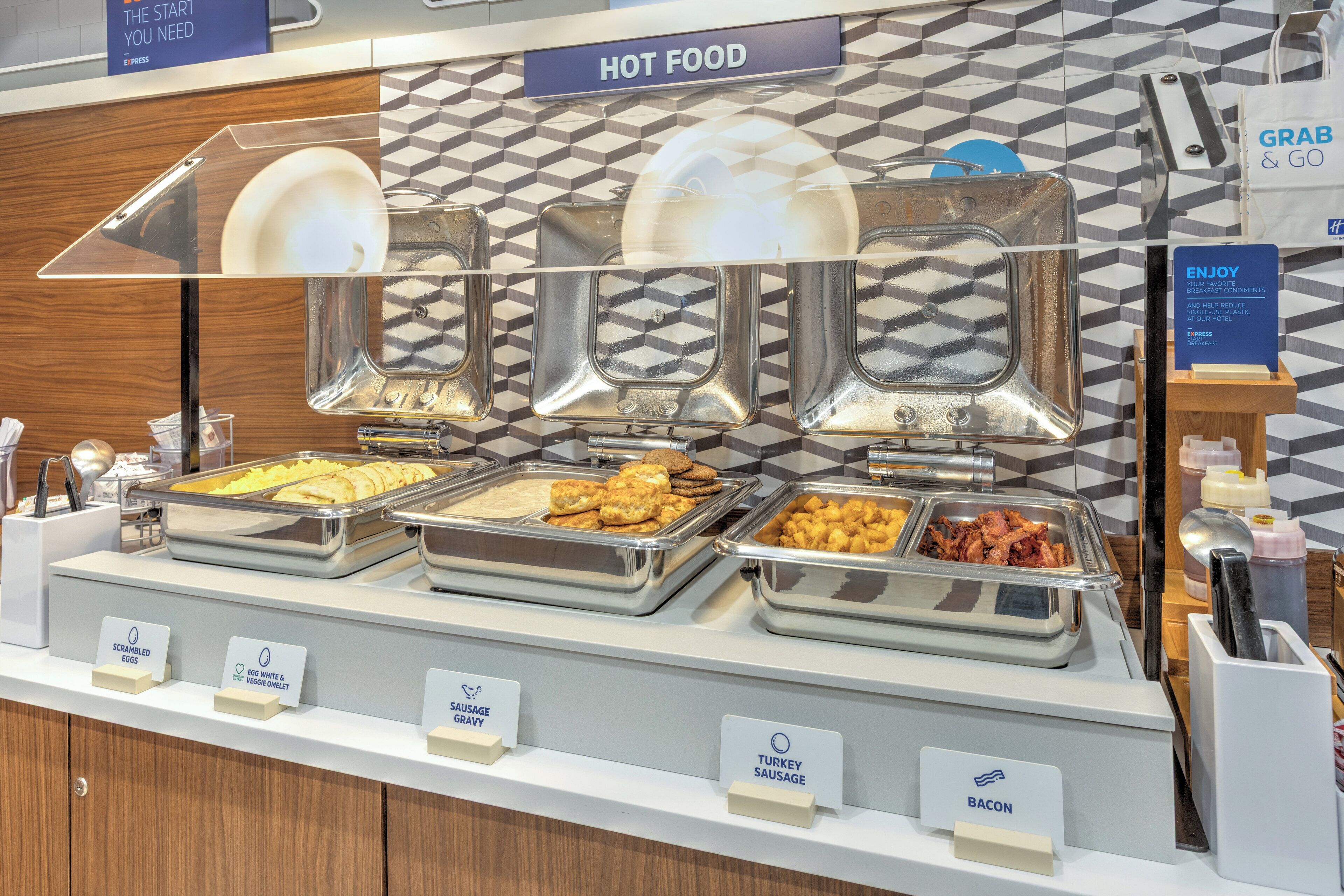 A buffet with labeled trays of scrambled eggs, white and brown cheese, sausage gravy, turkey sausage, and bacon.