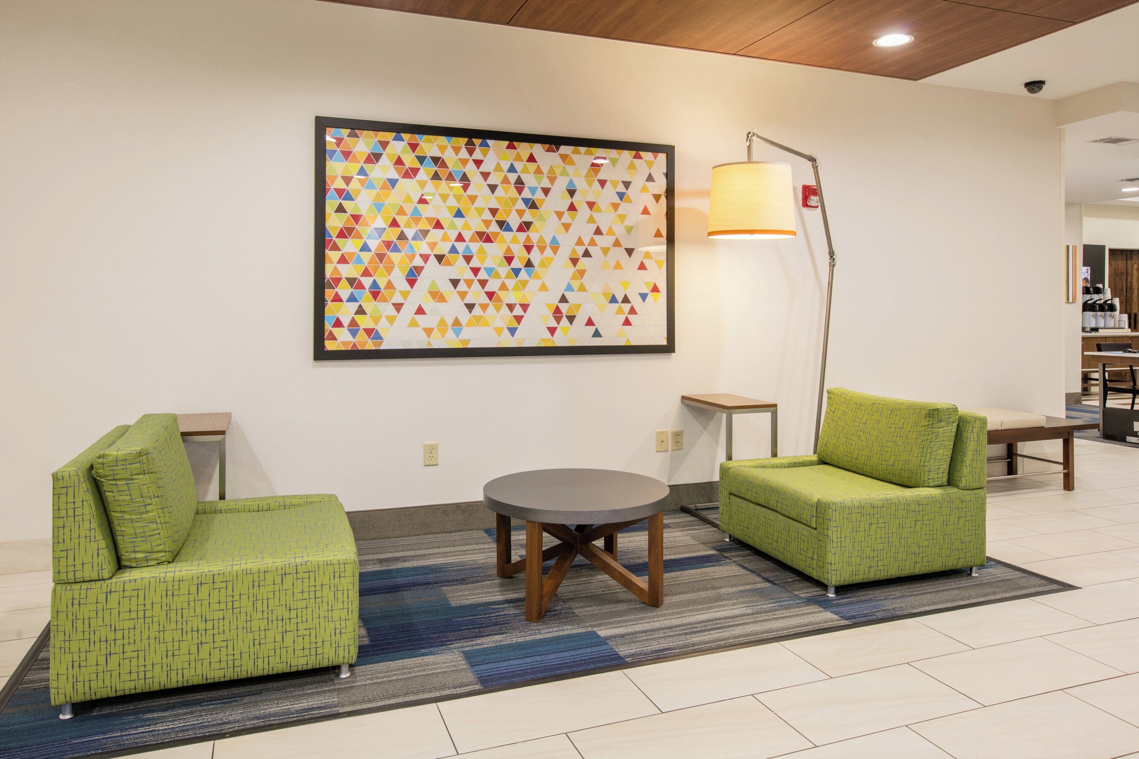 A modern lounge area with two green armchairs, a round table, and a colorful geometric wall art piece.