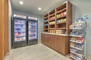 Een open zelfbedieningsgedeelte met gekoelde drankautomaten, diverse pakjes snacks, en een bord dat aangeeft dat de markt 24 uur per dag geopend is.
