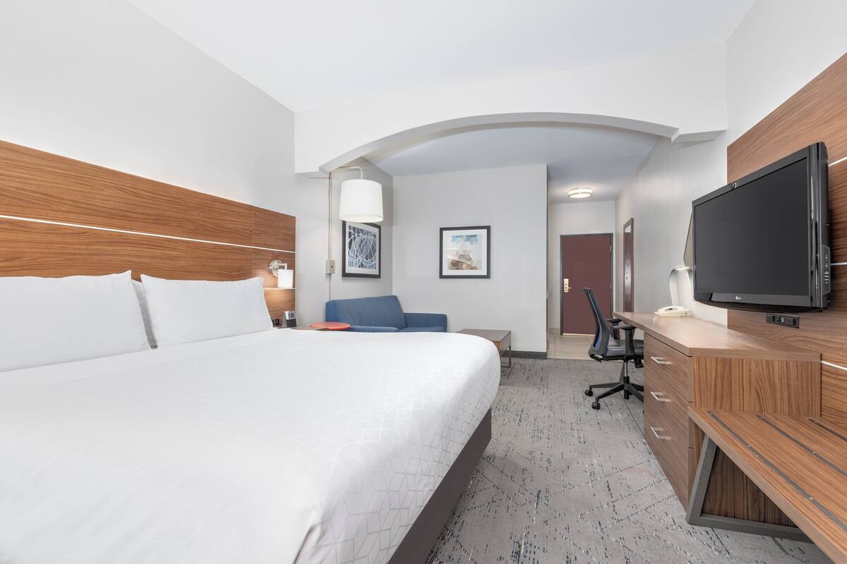 A hotel room with a large bed, a desk with a flat-screen TV, a blue chair, and a lamp.