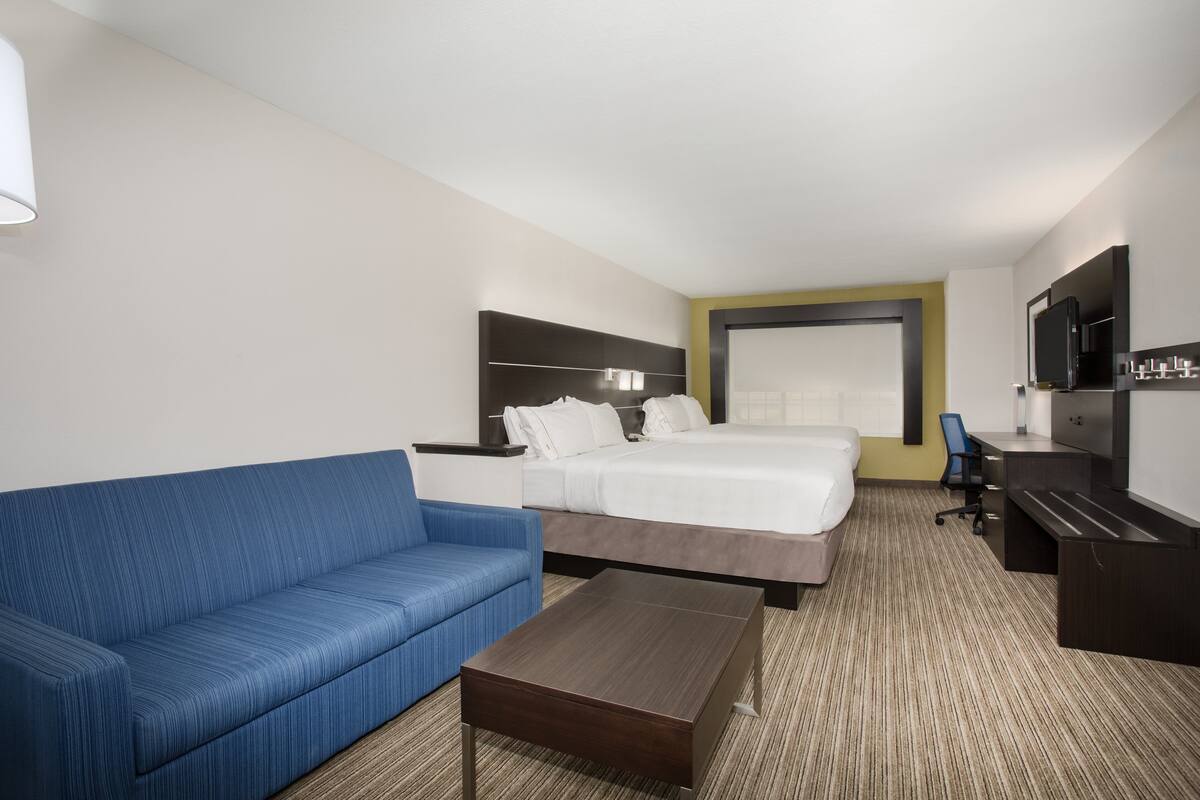 Quarto de hotel com uma cama, um sofá azul, uma mesa de centro de madeira, uma escrivaninha com cadeira e uma televisão fixada na parede.