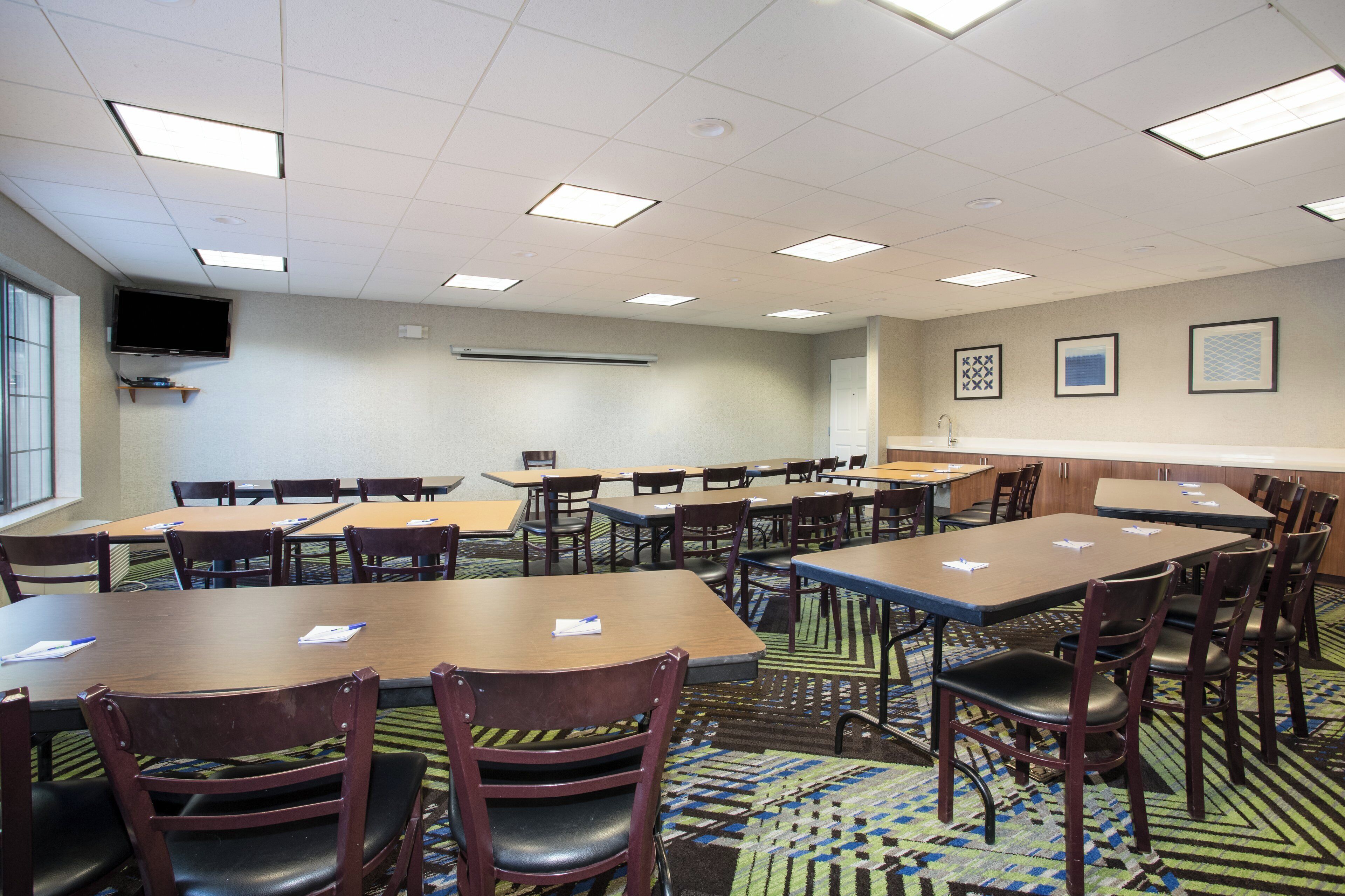 Une salle de conférence équipée de plusieurs tables et chaises, d’un téléviseur à écran plat fixé au mur et d’un tapis à motifs.