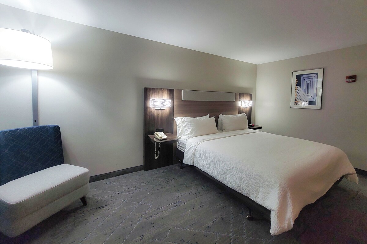 A hotel room with a bed, bedside table, lamp, chair, and artwork on the wall.