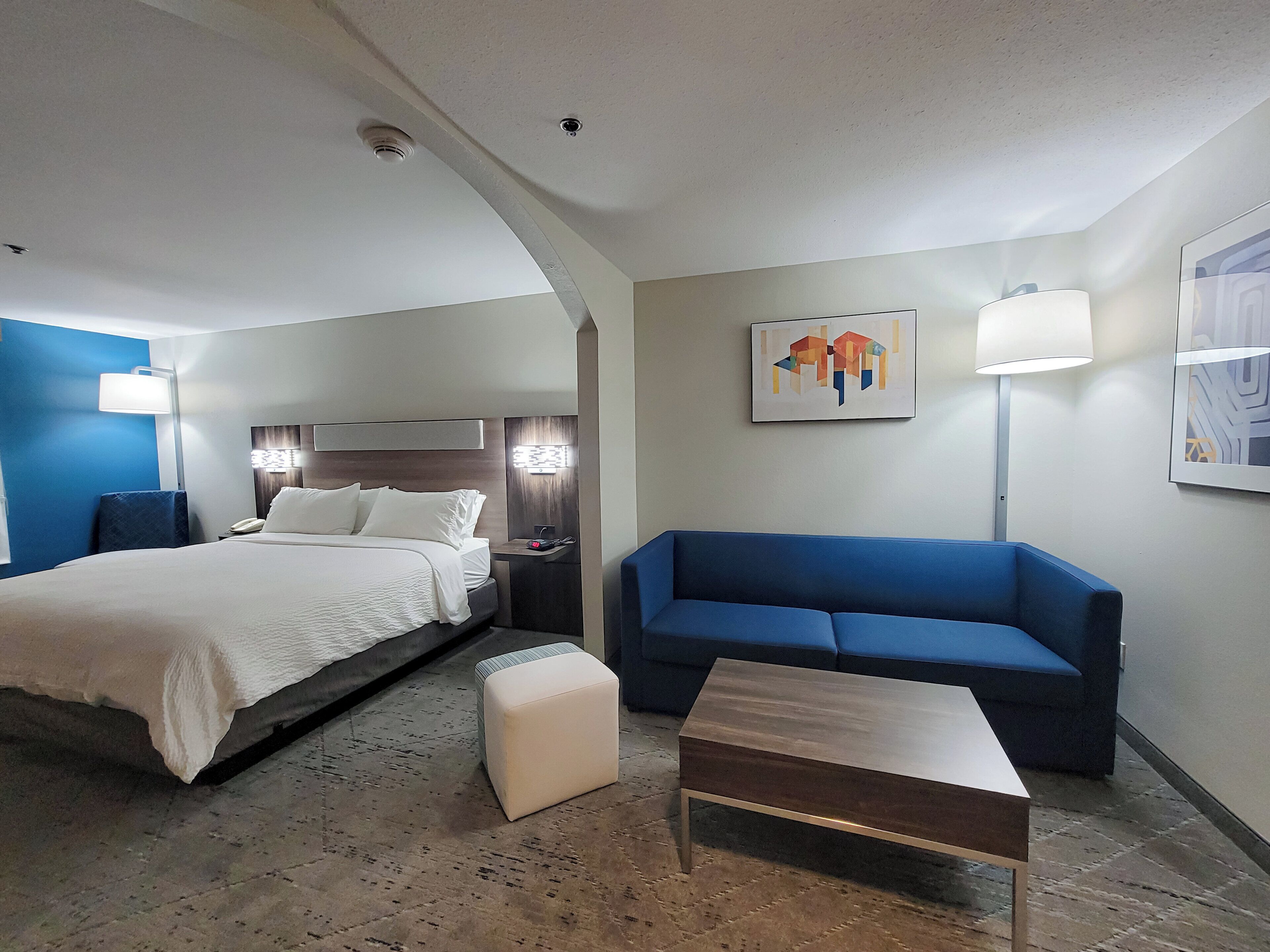 A hotel room with a bed, a blue sofa, a small table, and a nightstand with a lamp.