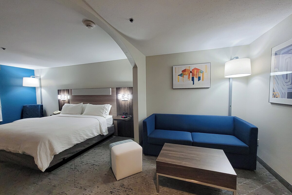 A hotel room with a bed, a blue sofa, a small table, and a nightstand with a lamp.