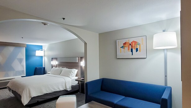 A modern hotel room with a bed, a blue sofa, a wooden coffee table, a lamp, and a painting on the wall. - Holiday Inn Express Hotel & Suites Enid - Highway 412 by IHG (Enid)