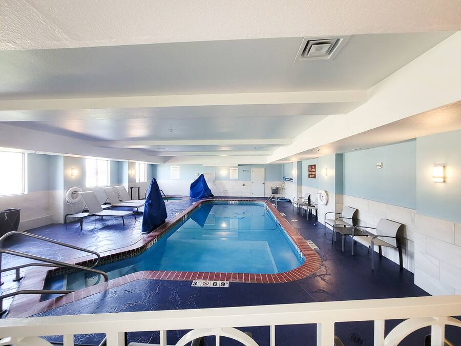 An indoor swimming pool with a blue tiled floor, white lounge chairs, and a poolside area with a small table and chairs.