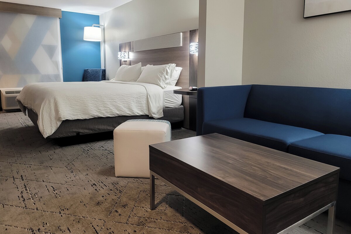 A modern hotel room with a bed, a blue sofa, a wooden coffee table, and a patterned rug.