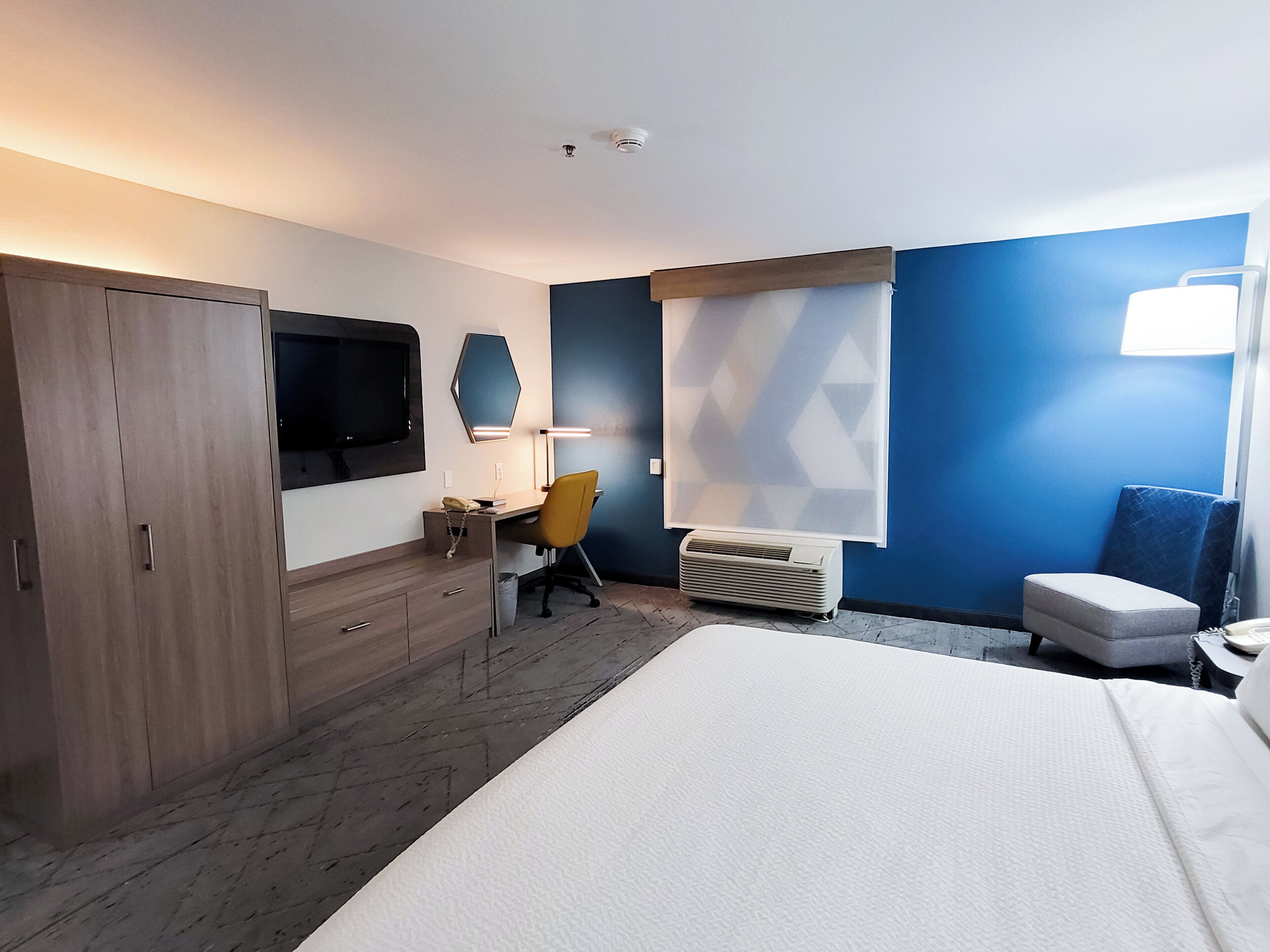 Habitación de hotel con cama, escritorio con silla, armario, televisor y ventana con persianas.