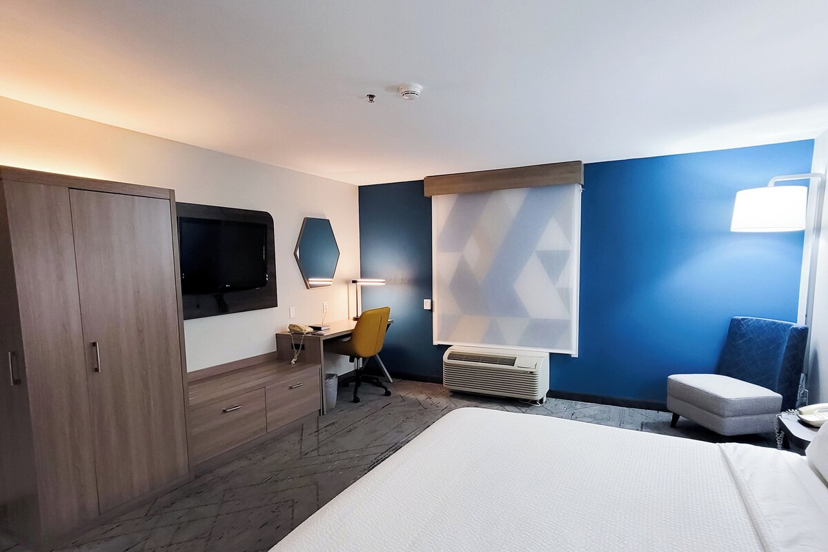 A hotel room with a bed, a desk with a chair, a TV, a mirror, a window with blinds, and a wardrobe.