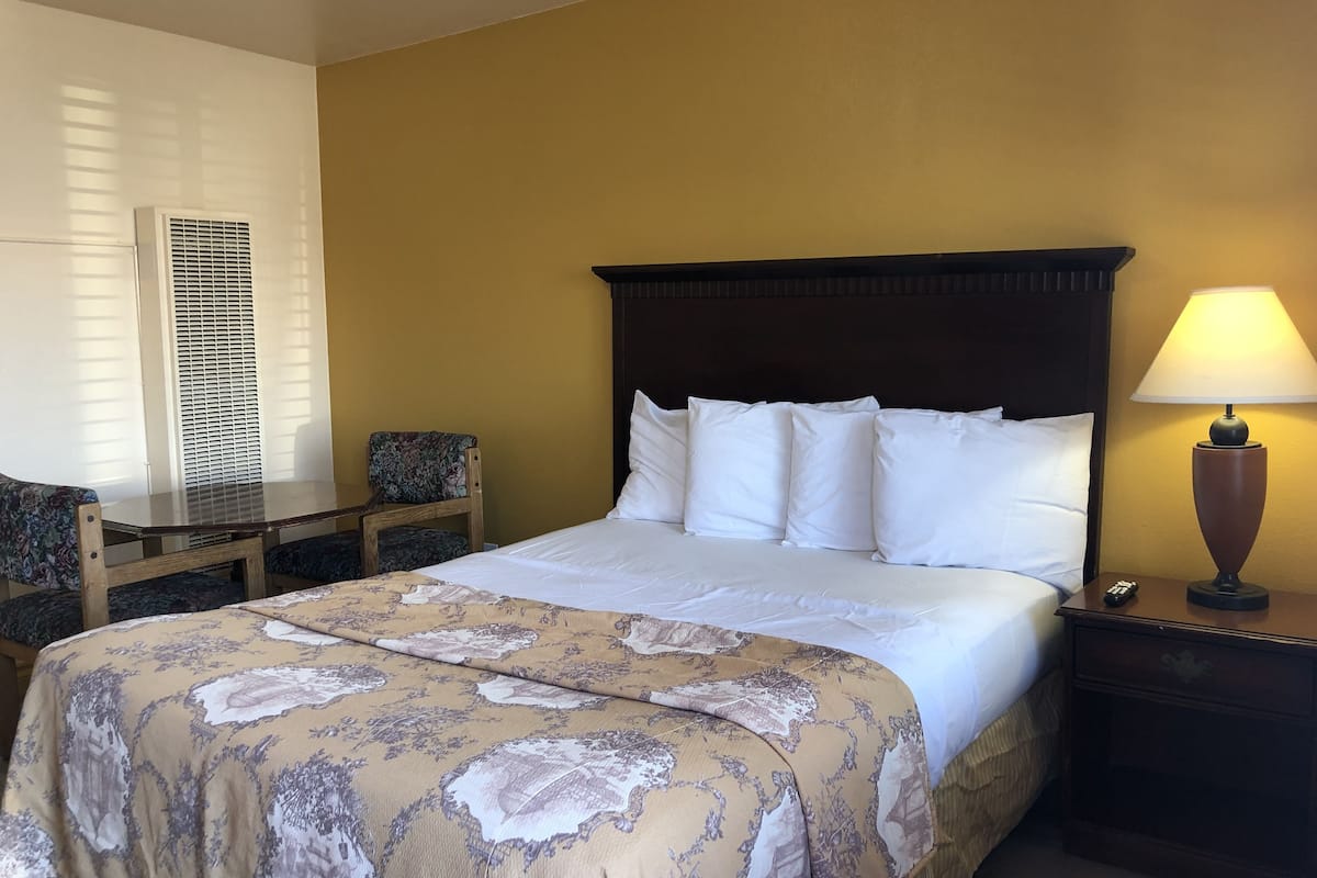 A hotel room with a bed, a nightstand, a lamp, a chair, and a window with blinds.