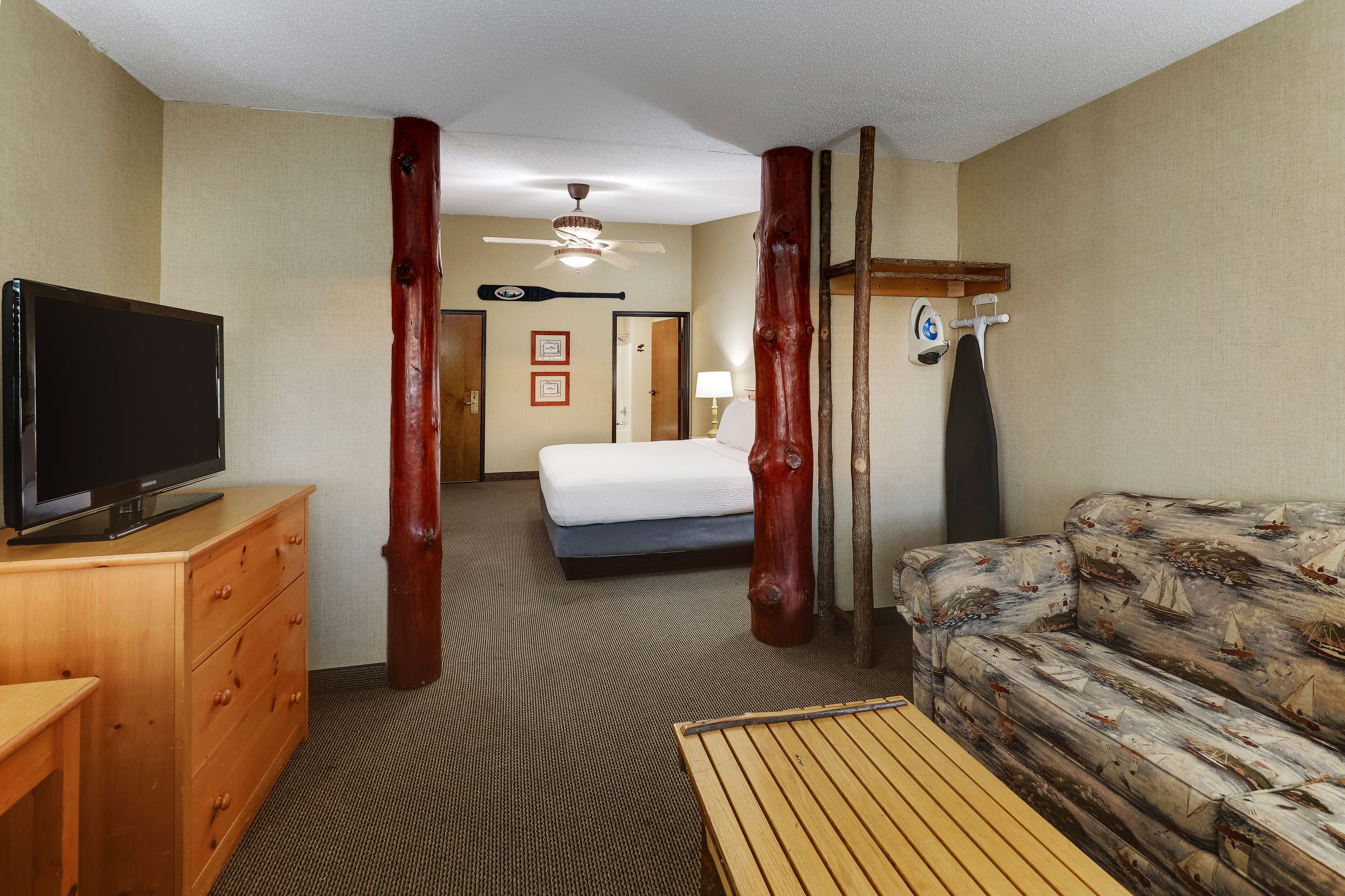 Habitación de hotel con cama, cómoda de madera, televisor, mesa de madera, sofá, ventilador de techo y puerta que da a otra habitación.