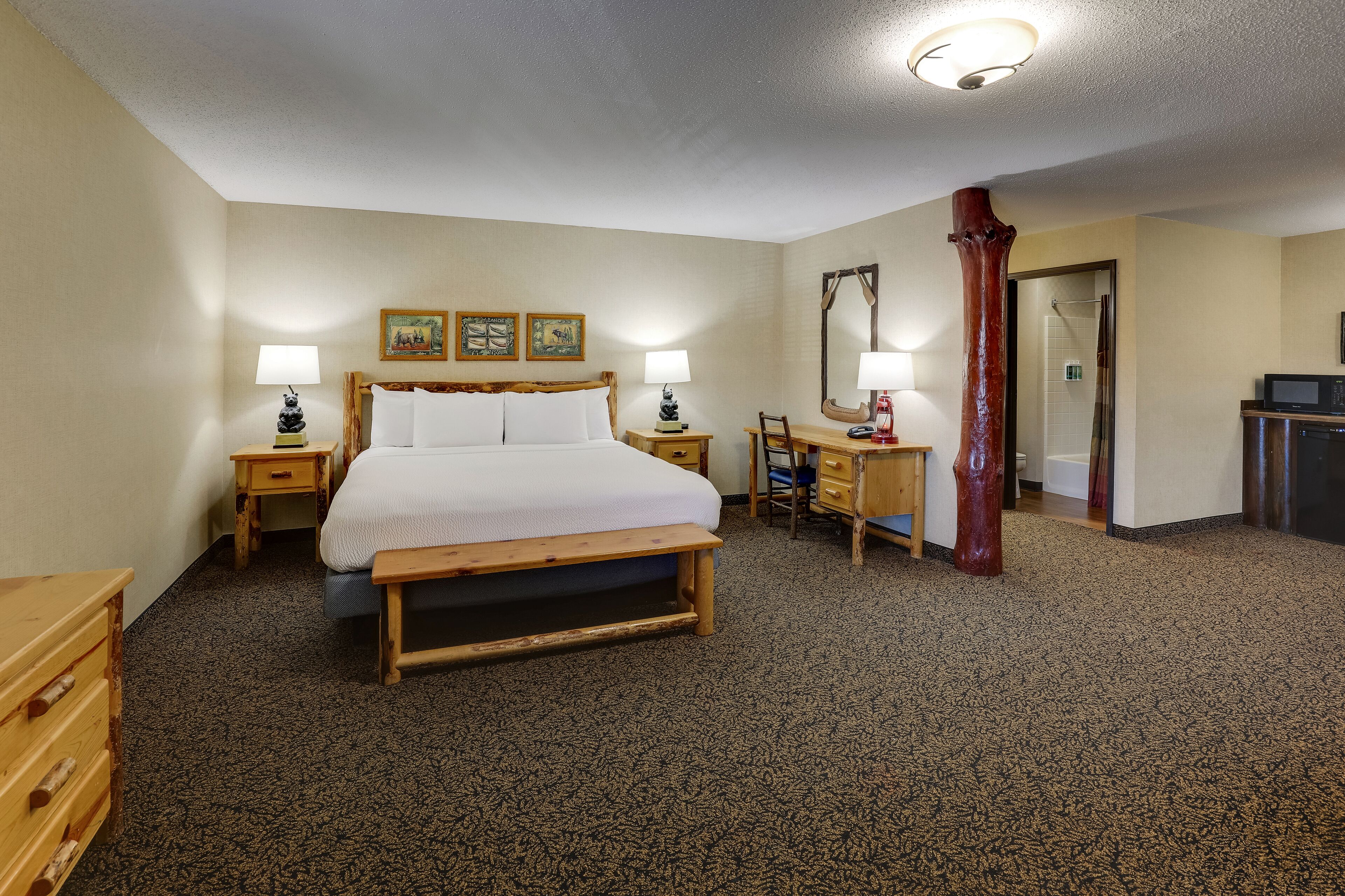 A hotel room with a large bed, two bedside tables with lamps, a wooden bench, a desk with a chair, a mirror, and a shower area.