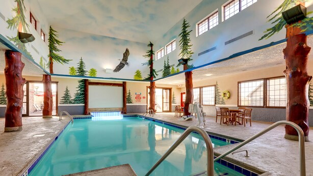 An indoor swimming pool with wooden pillars, a ceiling mural of trees and a bird, and a seating area with tables and chairs.