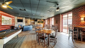 A spacious indoor area with a pool table, wooden tables, and chairs, featuring a brick wall and large windows.