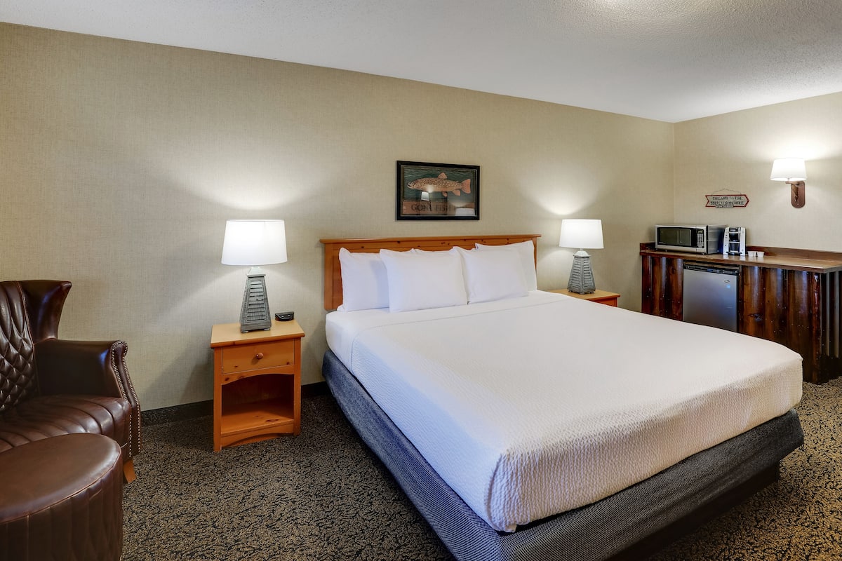 Habitación de hotel con una cama grande, dos mesitas de noche con lámparas, una mesita auxiliar de madera, un sillón de cuero marrón y un microondas sobre un mueble de madera.