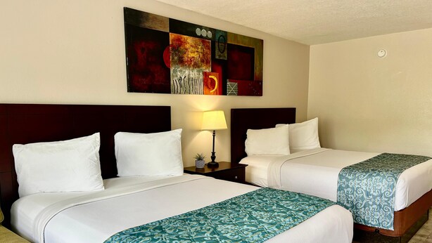 A hotel room with two beds, each with white linens and a patterned throw blanket. There is a nightstand with a lamp and a small plant. A large abstract painting is on the wall above the beds. - Shining Light Inn & Suites (Kissimmee)