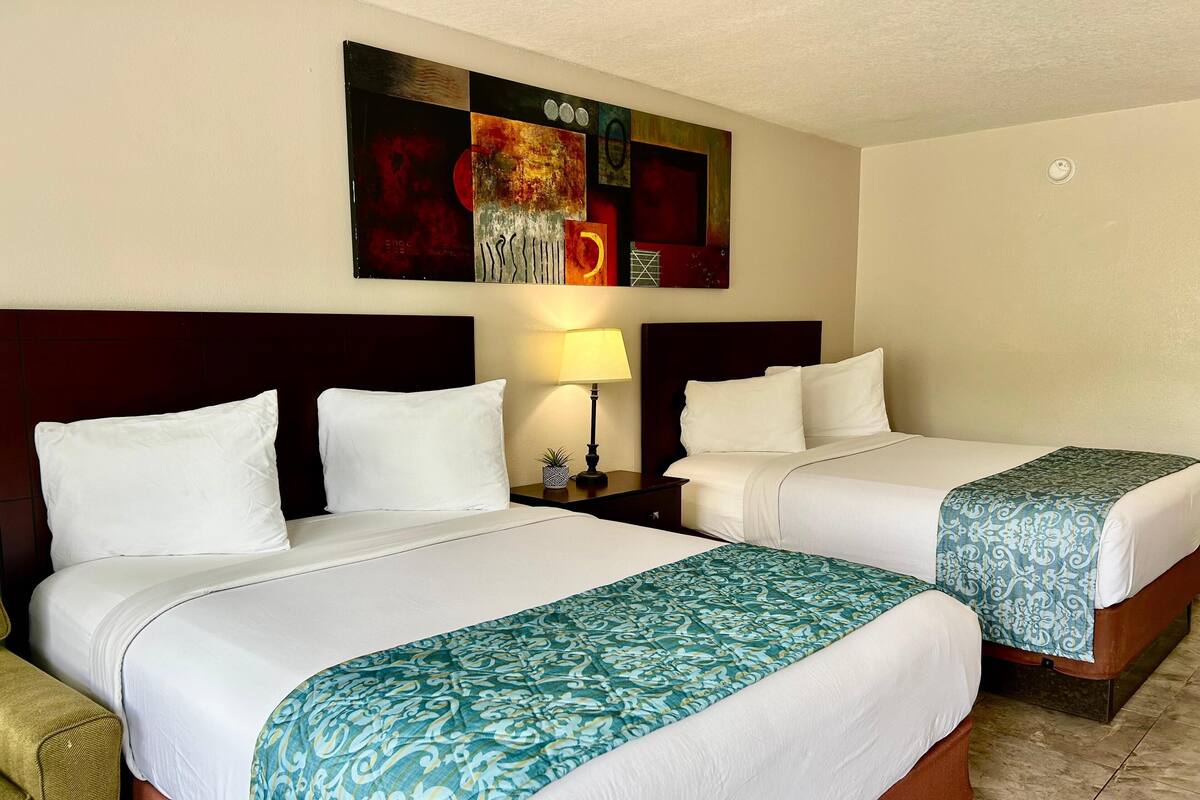 A hotel room with two beds, each with white linens and a patterned throw blanket. There is a nightstand with a lamp and a small plant. A large abstract painting is on the wall above the beds.