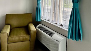 A room with a single window, teal curtains, a beige armchair, and a white air conditioning unit. - Shining Light Inn & Suites (Kissimmee)