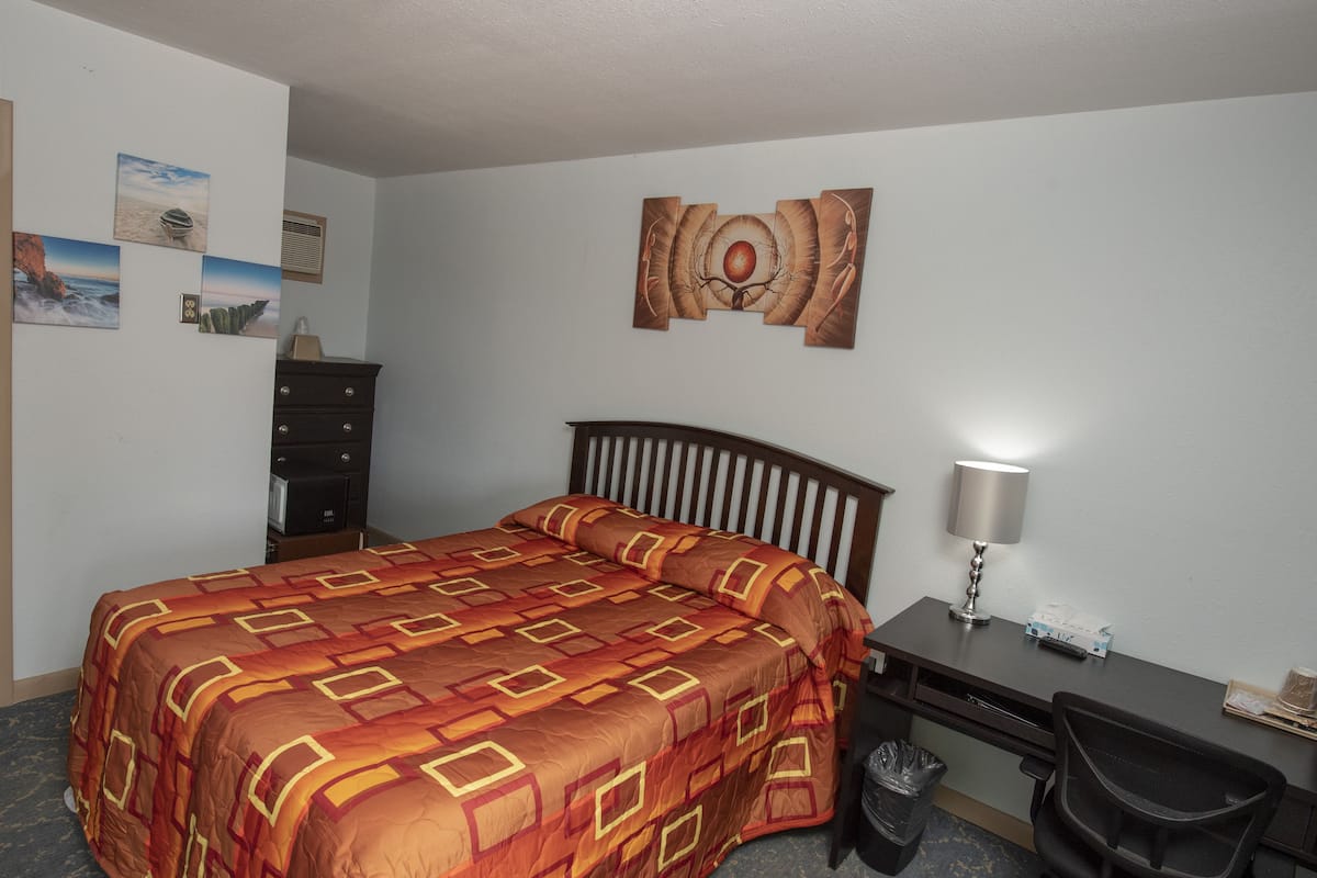 A hotel room with a bed, desk, chair, and a wall with artwork.
