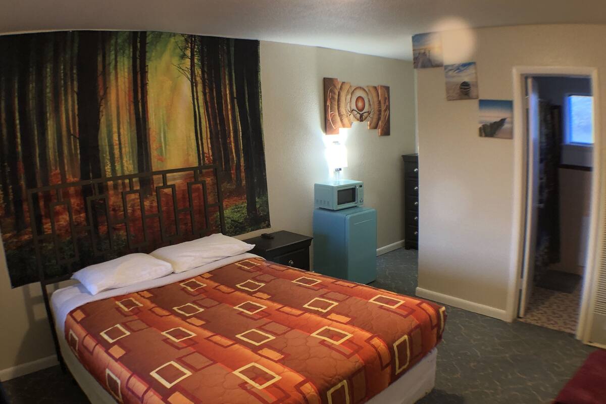 A hotel room with a bed, a nightstand, a small blue refrigerator, and a wall with a forest painting.