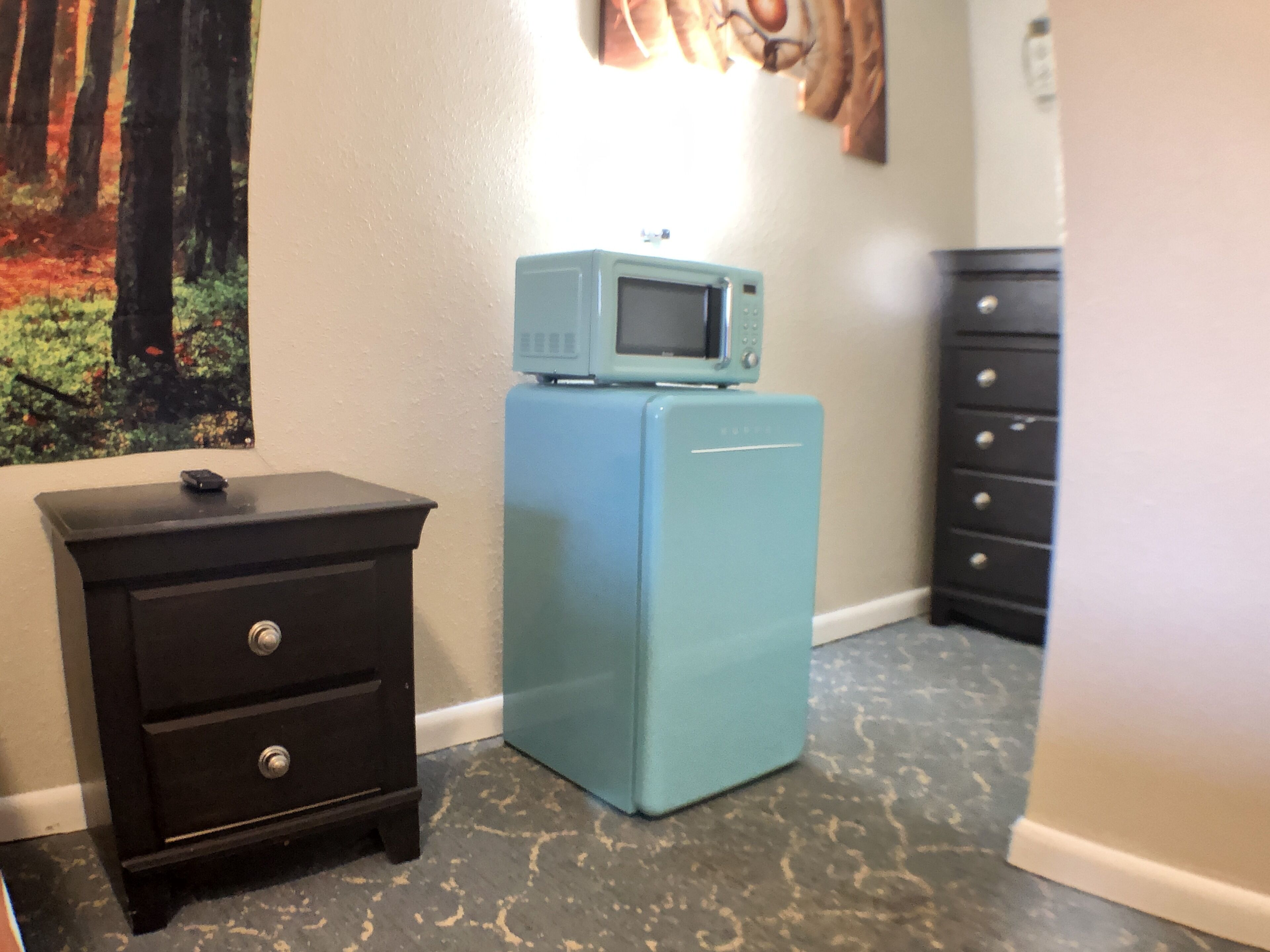 A room with a teal refrigerator, a black nightstand with drawers, and a wall-mounted TV.