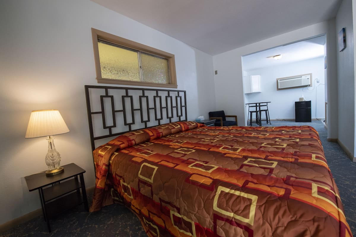 A hotel room with a large bed, a bedside table with a lamp, a window, and a kitchenette area.