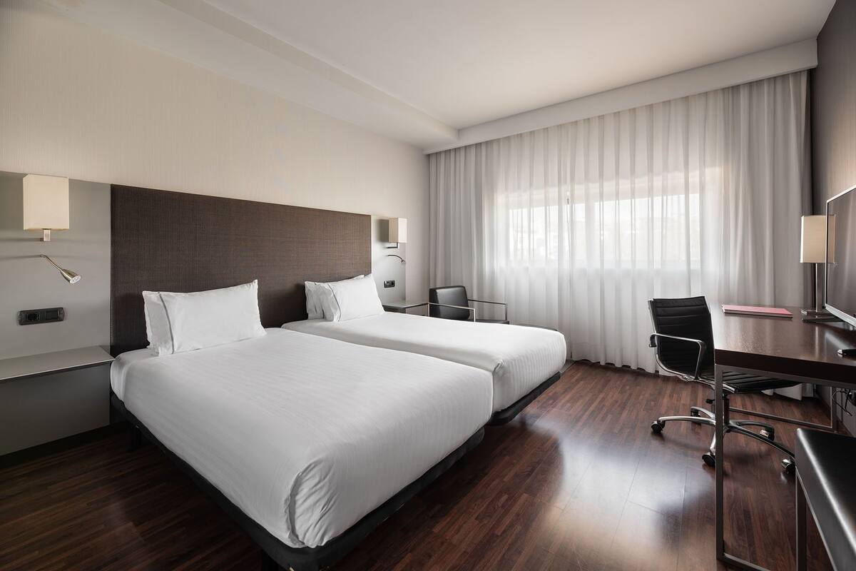 Una habitación de hotel con dos camas, un escritorio, una silla, un televisor y una ventana con cortinas.