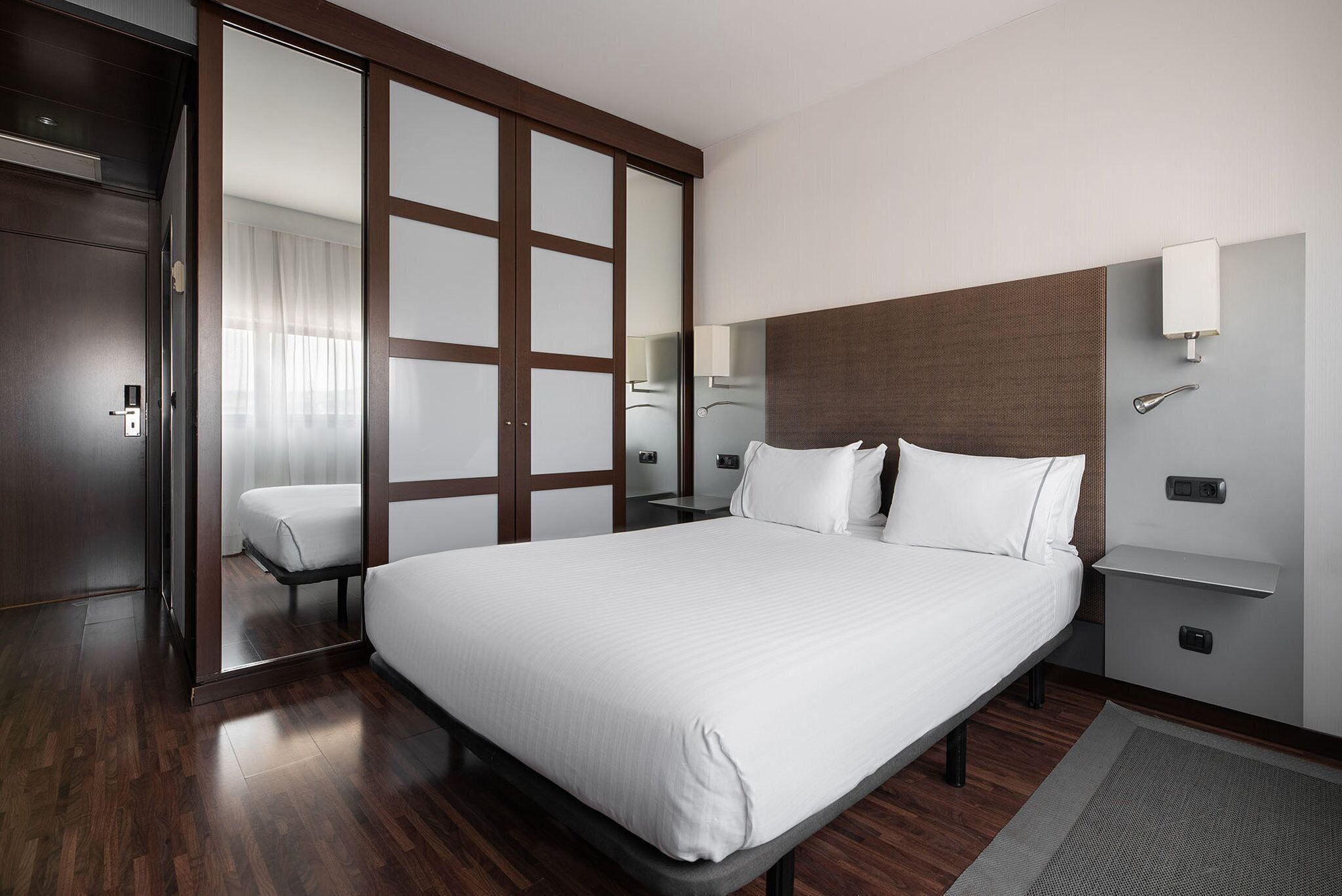Una habitación de hotel moderna con una cama grande, suelo de madera, puerta corredera, una mesita de noche y un tocador.