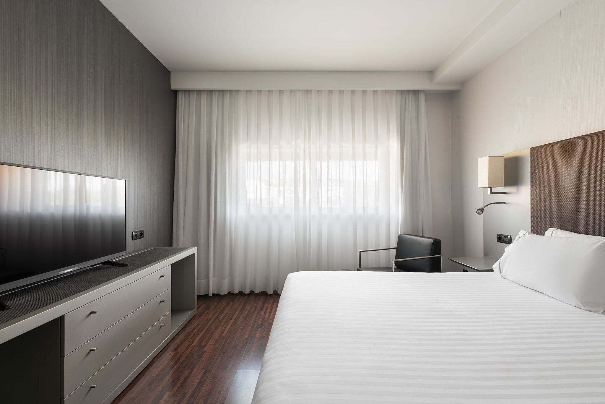 Una camera d'hotel moderna con un letto grande, una TV a schermo piatto su un mobile integrato, una scrivania con sedia e un'ampia finestra con tende leggere.