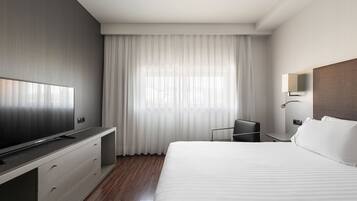 Una camera d'hotel moderna con un letto grande, una TV a schermo piatto su un mobile integrato, una scrivania con sedia e un'ampia finestra con tende leggere.