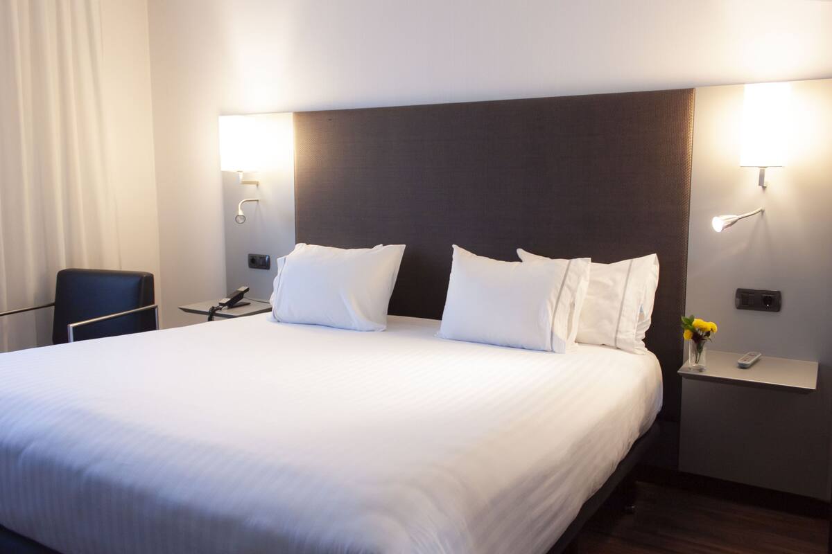 Habitación de hotel con una cama grande, dos mesitas de noche, una silla y una mesita con un florero.