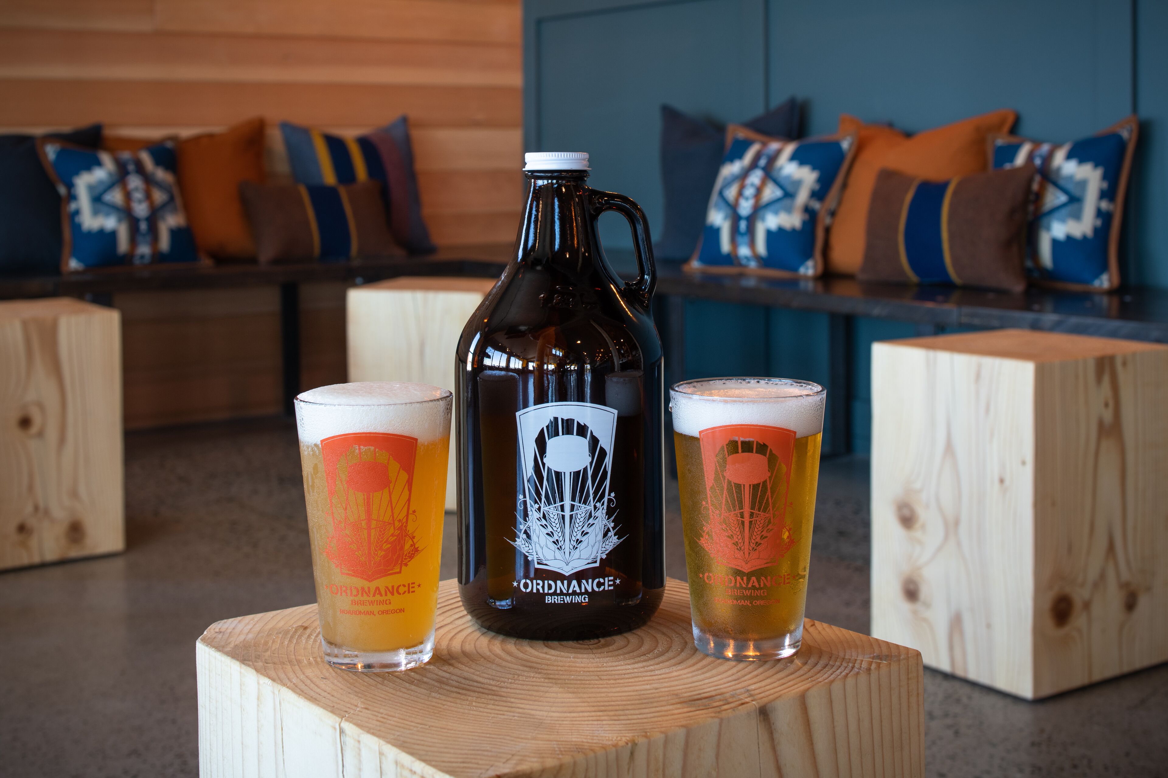 A wooden table with a dark glass carboy and two glasses of beer with 'ORDNANCE BREWING' branding.