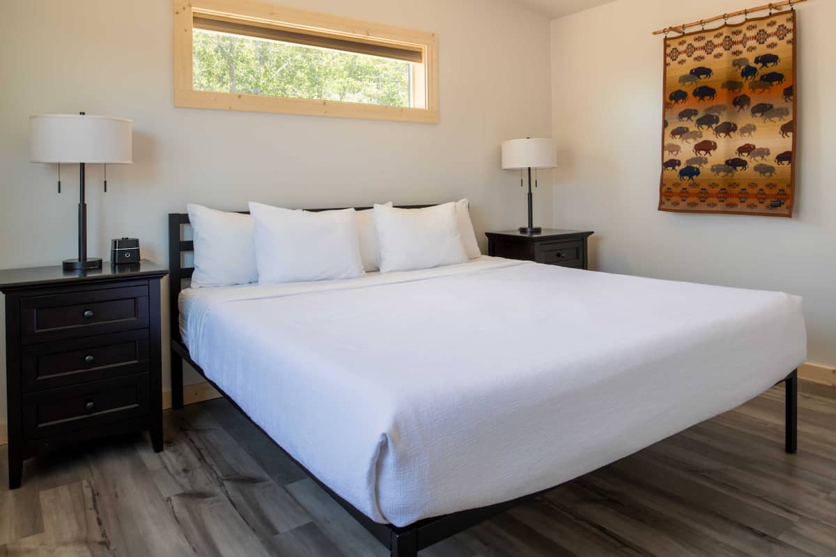 A neatly made bed with white linens, two bedside tables with lamps, a window with a wooden frame, and a patterned tapestry on the wall.