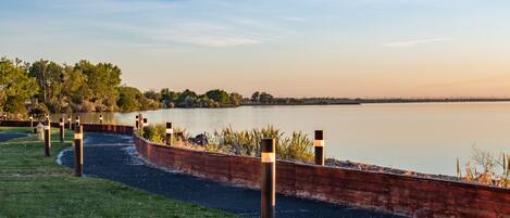 Un paseo junto al río con un muro de ladrillo y pilares, rodeado de césped y árboles.