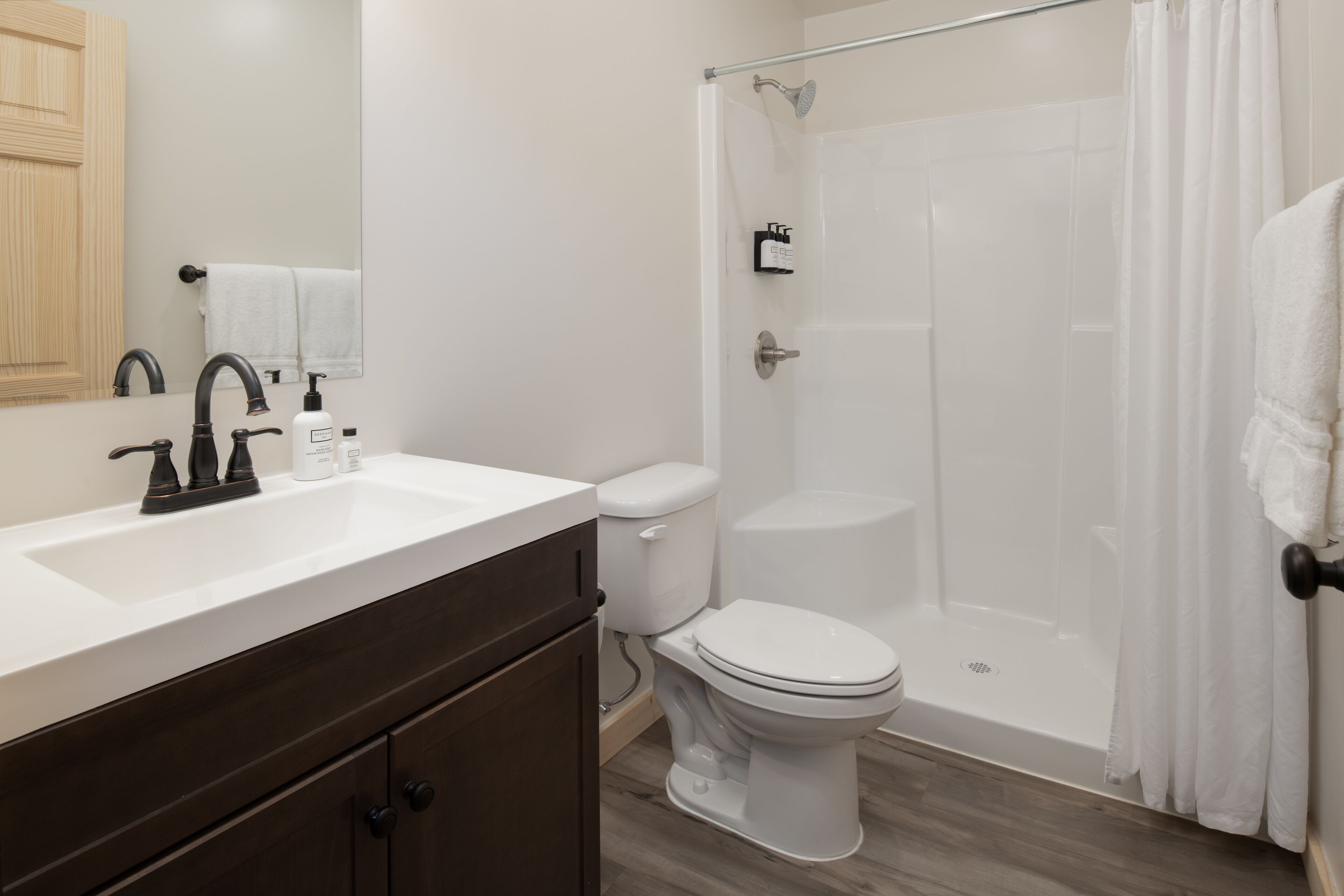 Un baño moderno con un lavamanos blanco, un mueble de madera oscura, un inodoro blanco y una zona de ducha con mampara de vidrio.