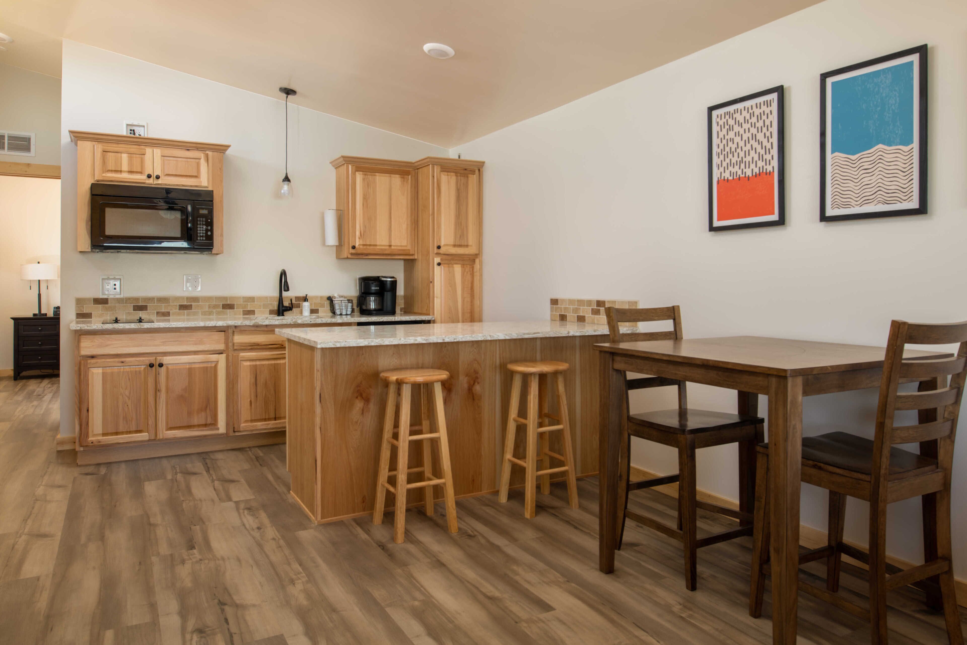Una cocina con armarios de madera, microondas, cafetera y un comedor con taburetes y una mesa.