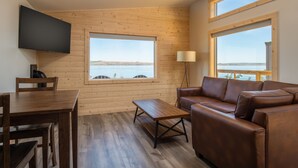A room with a wooden floor, a brown leather sofa, a wooden coffee table, a TV mounted on the wall, and large windows with a view of the water. - River Lodge and Cabins (Boardman)