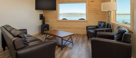 Uma sala de estar acolhedora com interior de madeira, sofás de couro, uma TV de tela plana e uma grande janela com vista para um lago.