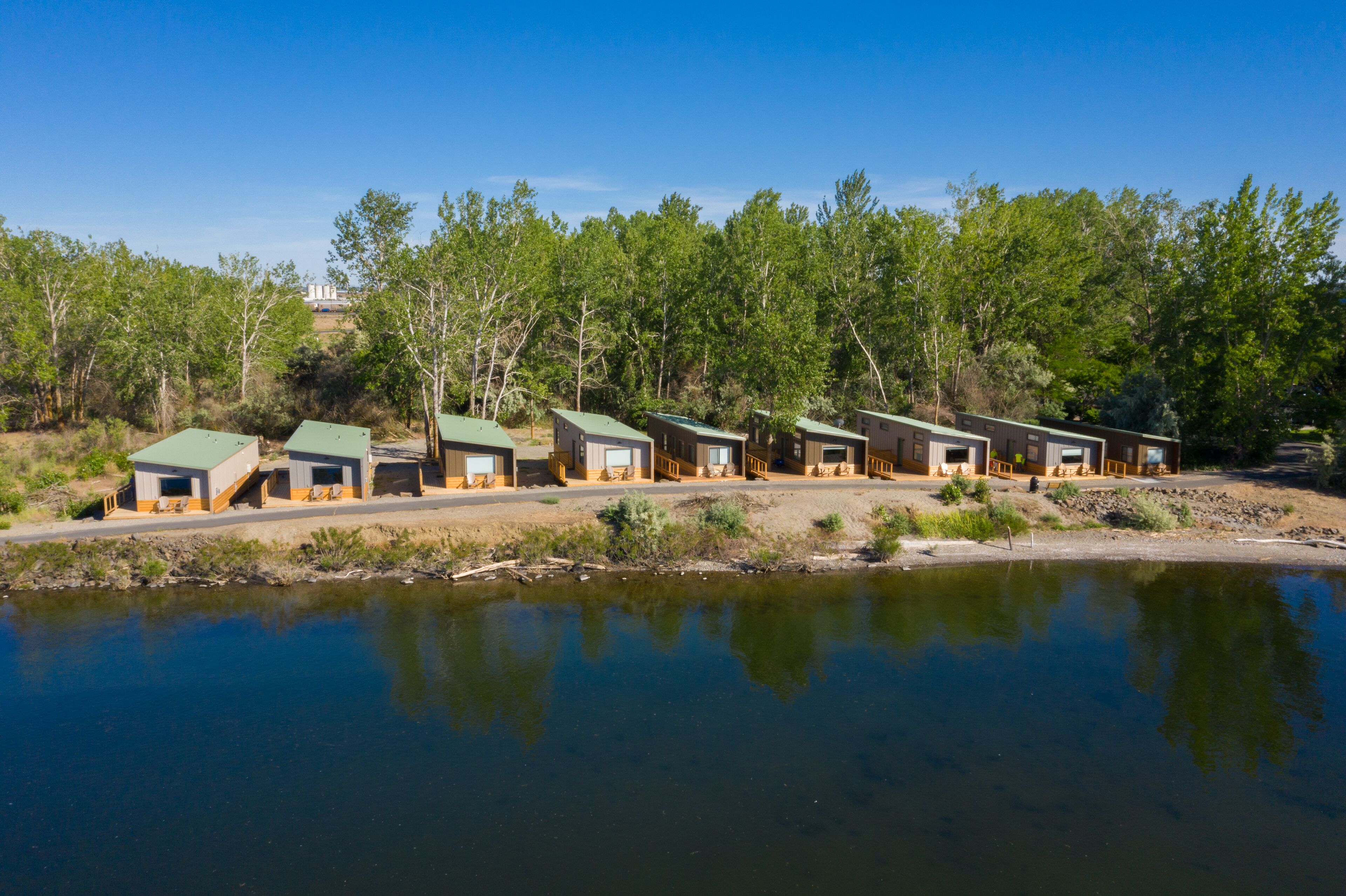 Une rangée de cabanes aux toits verts, situées au bord d’un lac paisible, entourées d’une forêt dense.