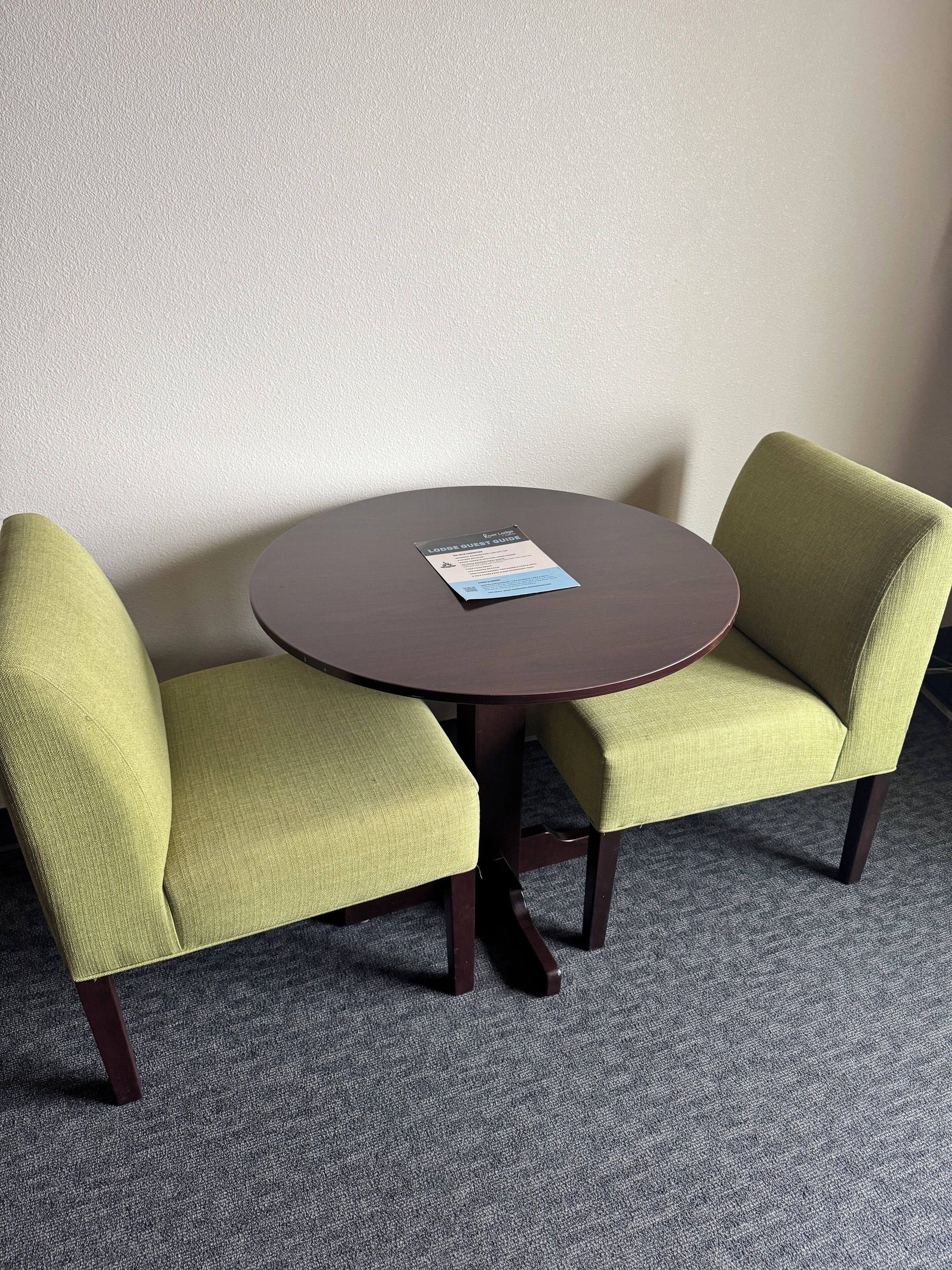 A small round table with two green upholstered chairs, a printed guide on the table, and a carpeted floor.