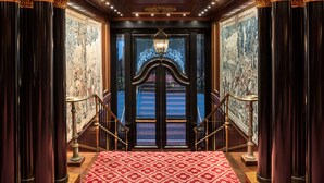 Une entrée imposante, ornée de colonnes richement décorées, un tapis rouge à motifs et une grande tapisserie aux détails complexes qui recouvre les murs.