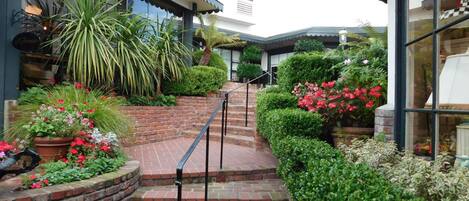 A brick pathway leading to a building with a covered entrance and a garden with various plants and flowers.