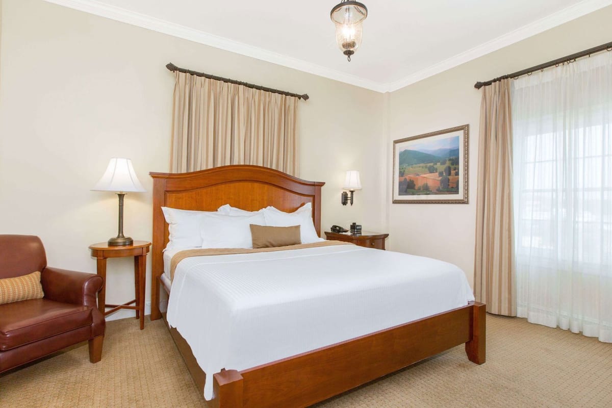 A hotel room with a large bed, a wooden headboard, two bedside tables with lamps, a leather armchair, and a framed landscape painting on the wall.